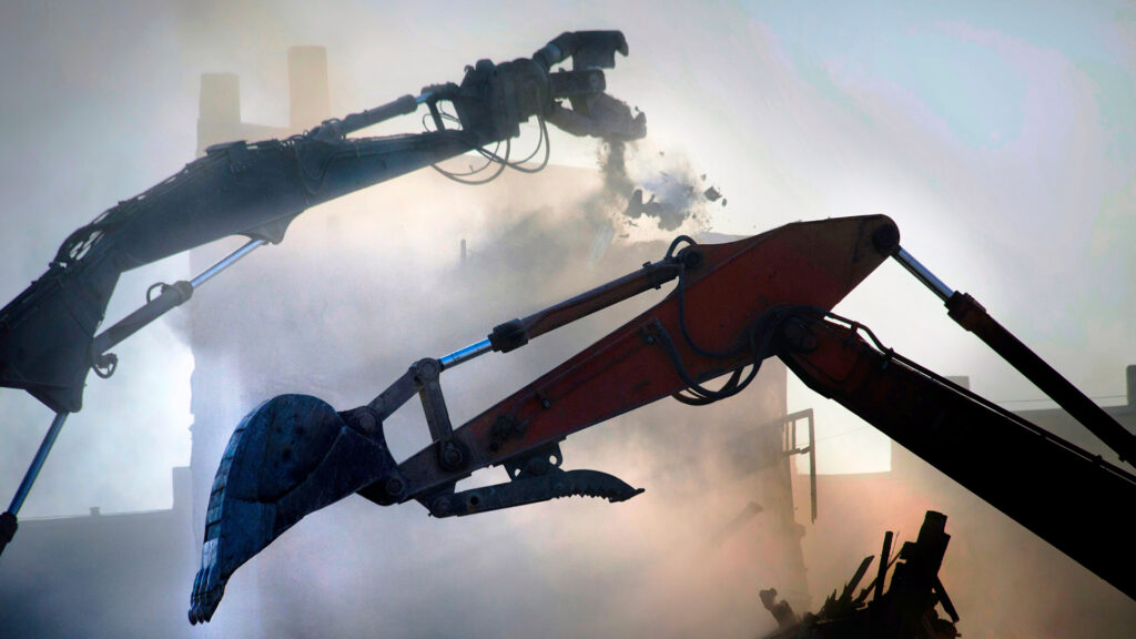 Excavators demolishing a structure with dust and debris in a backlit scene