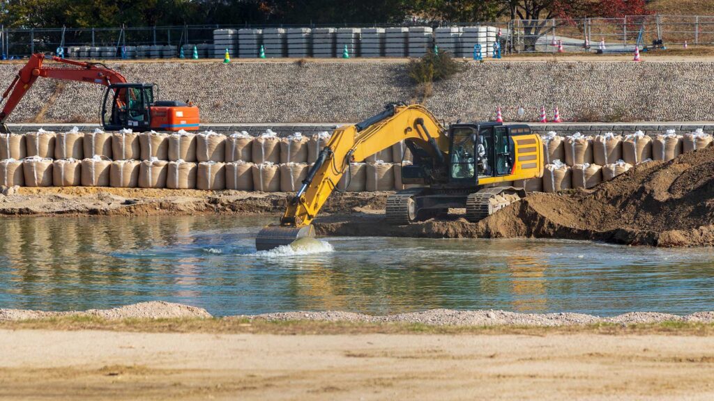 Excavator dredging water along a shoreline with erosion control barriers in place
