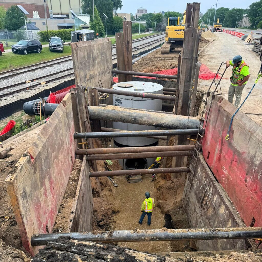Workers installing a large concrete utility structure in a trench near railroad tracks